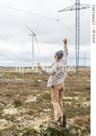 Woman in beige pants and a gray jacket raises her arms joyfully near wind turbines and power lines in Teriberka, Russia Woman in beige pants and a gray jacket raises her arms joyfully near wind turbines and power lines in Teriberka, Russia 130461962