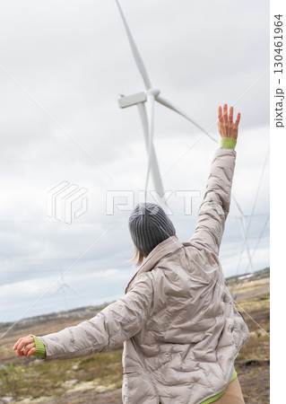 Woman in a gray jacket and hat raises her arms toward a large wind turbine in Teriberka, Russia 130461964