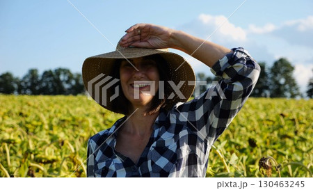 Happy smiling female farmer looks into camera standing near sunflower field. Portrait of adult beautiful agronomist in straw hat with yellow plantation at background. Agricultural business concept 130463245