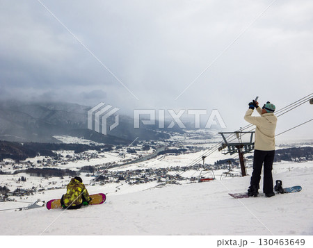 曇り空のスキー場でスマートフォンで雪景色を撮影するスノーボーダー (長野県、飯山市、戸狩温泉) 130463649