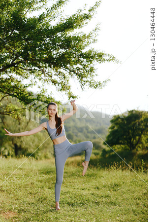 Young athletic woman practicing yoga outdoors in a serene green landscape, embodying tranquility and strength with a smile Young athletic woman practicing yoga outdoors in a serene green landscape, embodying tranquility and strength with a smile 130464448