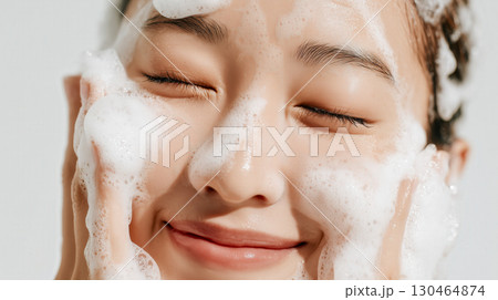 白背景のスタジオ撮影、泡立つ洗顔を楽しむ若い女性の顔アップ—清潔感とリラックスを感じる美肌ケアシーン 130464874