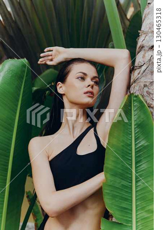 Woman in a black bikini top standing next to a palm tree with green leaves on a sunny tropical beach Woman in a black bikini top standing next to a palm tree with green leaves on a sunny tropical beach 130465318
