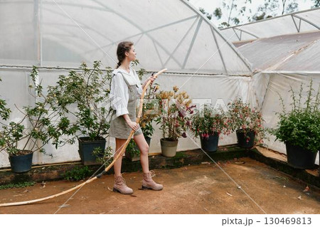 Young woman gardening in a greenhouse, wearing a casual outfit and boots, surrounded by blooming plants and flowers, embodying a nurturing and vibrant atmosphere 130469813