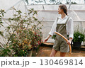 Young woman in a greenhouse wearing a white shirt and green overalls, tending to plants with a hose, expressing joy and connection to nature 130469814