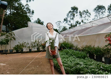 Young woman holding a potted plant in a greenhouse, showcasing joy and connection to nature with a casual outfit and lush greenery in the background 130469815