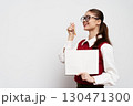Smiling young woman holding a blank notebook and pencil, wearing glasses, dressed in a stylish red vest against a neutral background, representing creativity and education 130471300