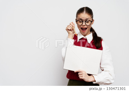 Happy girl in glasses holding a blank sign, smiling and pointing at it with a pencil, wearing a red sweater, on a plain white background Engaging and cheerful expression 130471301