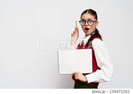 surprised girl with open notebook and pen, wearing glasses, standing against a light gray background, dressed in a red vest and white shirt, showing excitement in her expression 130471302