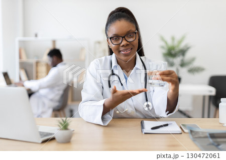 Female African doctor wearing white coat holding glass of water, discussing hydration benefits during consultation in bright office with professional environment 130472061