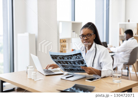 African female doctor analyzing patient x-rays in a modern office with natural light. Healthcare professional demonstrating concentration and expertise African female doctor analyzing patient x-rays in a modern office with natural light. Healthcare professional demonstrating concentration and expertise 130472071