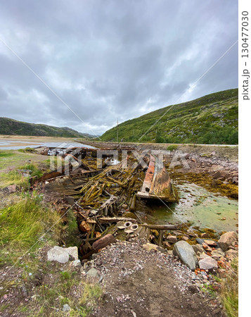 Decaying wooden shipwreck remains on rocky shore under cloudy mountain sky. 130477030