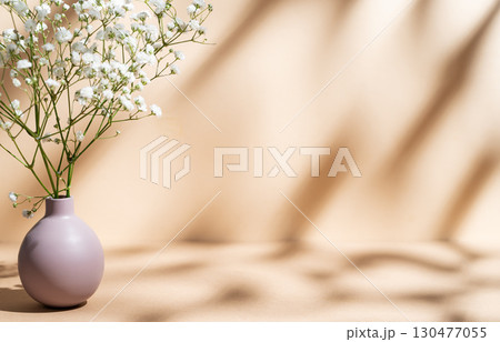 Delicate white gypsophila flowers in a lilac vase on a pastel beige background with sunny shadows. 130477055