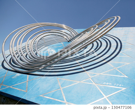 Contemporary metal sculpture on a vibrant blue wall in an urban outdoor setting under clear skies 130477106