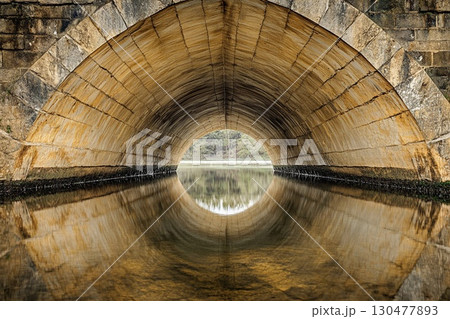 Vaulted bridge structure creates stunning mirror reflection beneath in calm water during early morning light 130477893