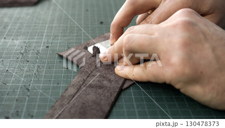 Close up of artisan thinning brown leather strip with sharp blade tool on green cutting mat 130478373