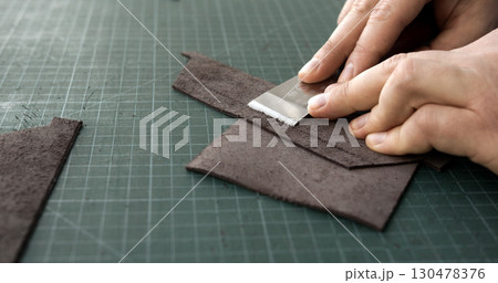 Leatherworker skiving leather piece with blade tool on green cutting mat 130478376