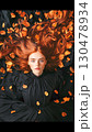 Beautiful redheaded woman with freckles wearing black dress, reclining against black backdrop scattered with vibrant orange autumn leaves 130478934