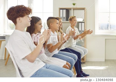Happy children sitting in a row on chairs and clapping hands at a school event in classroom Happy children sitting in a row on chairs and clapping hands at a school event in classroom 130484098