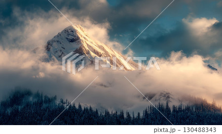 A mountain covered in clouds with a bright sun shining on it. The sky is blue and the clouds are white 130488343