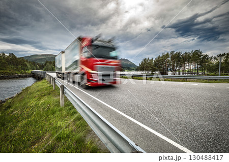 Red semi truck is driving down a road with a cloudy sky in the background. Red semi truck is driving down a road with a cloudy sky in the background. 130488417