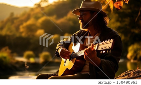 Mature guitarist wearing hat and glasses, strumming acoustic instrument near riverbank during warm sunset lighting 130490963