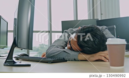 Exhausted Asian professional resting head on desk, glasses askew, surrounded by keyboard, mouse, and empty coffee cup during late night work session 130490992