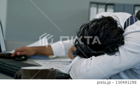 Tired businessman napping at cluttered desk, head resting on paperwork. Professional overwhelmed by work stress, seeking momentary rest during intense corporate environment 130491298