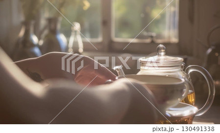 Woman serving blooming tea in transparent teapot, sunlight illuminating warm interior setting near window, creating peaceful afternoon mood 130491338
