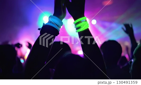 Concert crowd waving glowstick bracelets, cheering passionately during live music performance, sharing collective excitement and energy under vibrant stage lighting 130491359