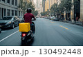 Delivery driver riding scooter on busy city street, delivering food or parcel with yellow insulated backpack on a sunny summer day 130492448