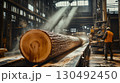 Inside a sawmill, a large tree trunk moves along a conveyor belt while a carpenter supervises the process, ensuring efficient wood production 130492450