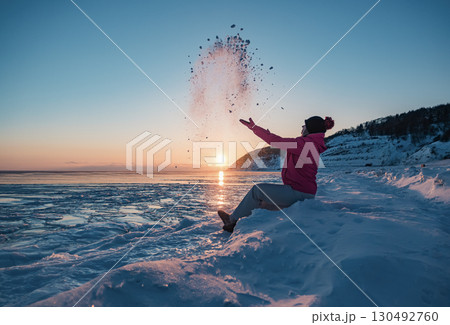 A young woman enjoying a winter sunset on the frozen shore while playfully tossing snow into the air A young woman enjoying a winter sunset on the frozen shore while playfully tossing snow into the air 130492760