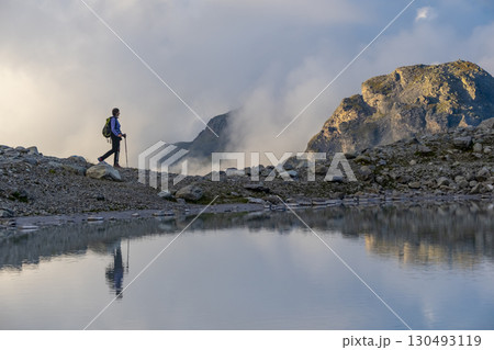 A person stands on rocky terrain, gazing at a stunning turquoise mountain lake surrounded. Dressed in hiking gear and holding trekking poles. Wildsee Pizol, Switzerland. A person stands on rocky terrain, gazing at a stunning turquoise mountain lake surrounded. Dressed in hiking gear and holding trekking poles. Wildsee Pizol, Switzerland. 130493119