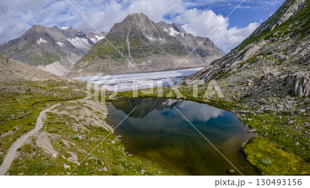 A stunning view of the Swiss Alps showcases a tranquil pond surrounded by rocky terrain, Aletsch Glacier and lush greenery, with towering mountains and glaciers in the background under a vibrant sky. 130493156