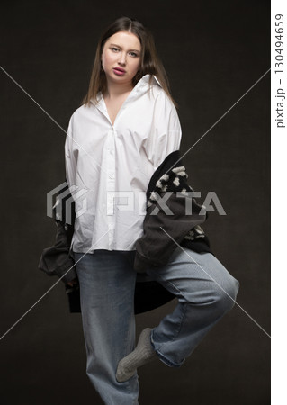 A beautiful girl in a white shirt and jeans stands on a gray background. 130494659