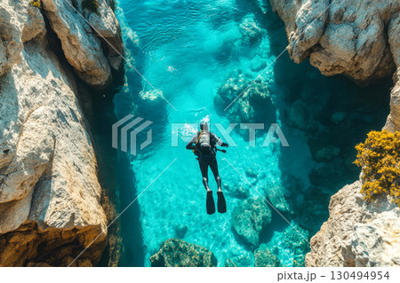 Scuba diver swimming through vibrant clear turquoise ocean water. Steep rugged rocky cliffs frame the beautiful underwater adventure and exploration. 130494954