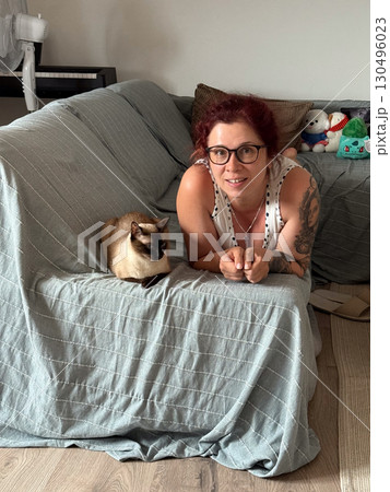 A woman with glasses and tattoos lies on a sofa next to her siamese cat. Home atmosphere, companionship, and lifestyle moments reflect connection, relaxation, and daily human-animal bond. 130496023