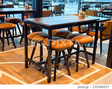 Tall wooden table with leather bar stools in a public cafe area. Representation of socialization, leisure, and hospitality through travel atmosphere and lifestyle dining culture. Tall wooden table with leather bar stools in a public cafe area. Representation of socialization, leisure, and hospitality through travel atmosphere and lifestyle dining culture. 130496057