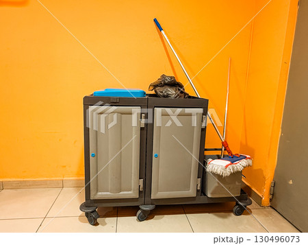 Cleaning cart with bins and mop against orange wall. Hygiene, maintenance, and sanitation support in public service environments. 130496073