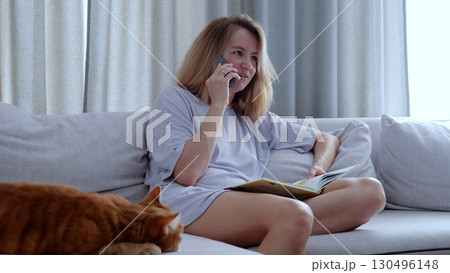 Young woman relaxes on a cozy sofa, chatting on her cellphone and enjoying a book while her ginger cat naps nearby 130496148