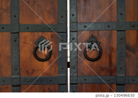 Close up of vintage wooden doors with iron reinforcements and intricate ring handles, symbolizing craftsmanship and strength. Classic design combining natural materials and historical elegance Close up of vintage wooden doors with iron reinforcements and intricate ring handles, symbolizing craftsmanship and strength. Classic design combining natural materials and historical elegance 130496630