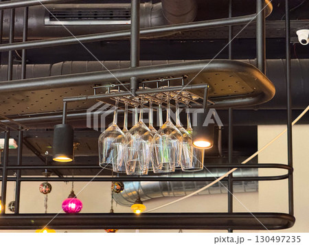 Wine glasses hanging upside down on ceiling rack in modern bar interior. Hospitality, catering, and lifestyle atmosphere combining drinkware, organization, and contemporary furnishing. Wine glasses hanging upside down on ceiling rack in modern bar interior. Hospitality, catering, and lifestyle atmosphere combining drinkware, organization, and contemporary furnishing. 130497235