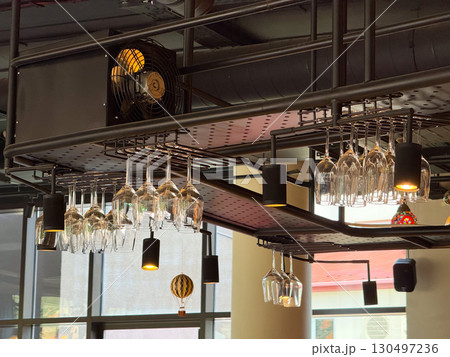 Wine glasses hanging upside down on ceiling rack in modern bar interior. Hospitality, catering, and lifestyle atmosphere combining drinkware, organization, and contemporary furnishing. Wine glasses hanging upside down on ceiling rack in modern bar interior. Hospitality, catering, and lifestyle atmosphere combining drinkware, organization, and contemporary furnishing. 130497236