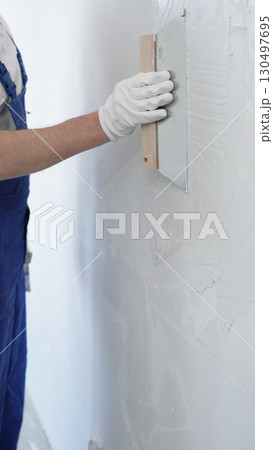 Male construction worker wearing protective gloves and blue construction coveralls, spreading plaster smoothly across wall using professional drywall taping knife during renovation, vertical view 130497695