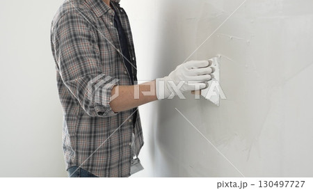 Unknown construction worker man wearing gloves and plaid shirt is using a putty knife to apply plaster on a white wall, during a home renovation or construction project 130497727