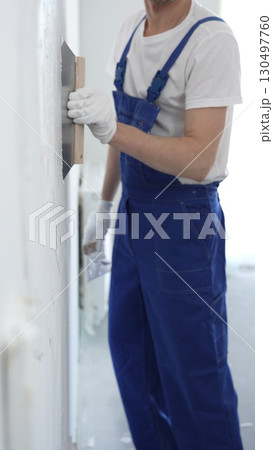 Man construction worker wearing protective gloves and blue construction coveralls, spreading plaster smoothly across wall using professional drywall taping knife Man construction worker wearing protective gloves and blue construction coveralls, spreading plaster smoothly across wall using professional drywall taping knife 130497760
