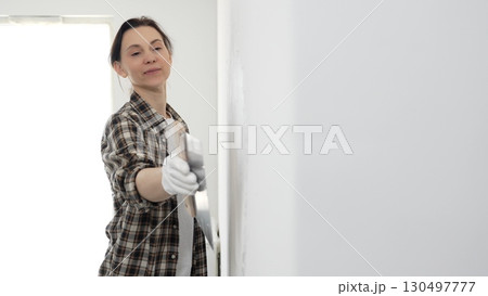 Female construction professional wearing checkered shirt is smoothing white wall surface, applying putty with professional spatula technique, showcasing skilled interior renovation work 130497777