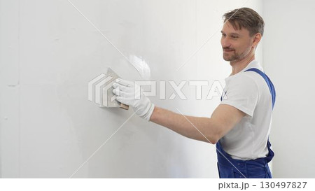 Middle aged man construction worker wearing gloves and blue overalls is using a putty knife to apply plaster on a white wall, performing home renovation and improvement 130497827