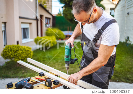 Mature male carpenter using electric drill on a wooden plank outdoors Mature male carpenter using electric drill on a wooden plank outdoors 130498587